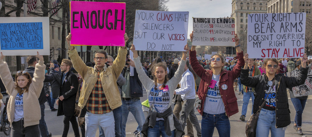 People protesting against gun violence. Signs have messages like "Enough" and "Your Guns Have Silencers But Our Voices Do Not"