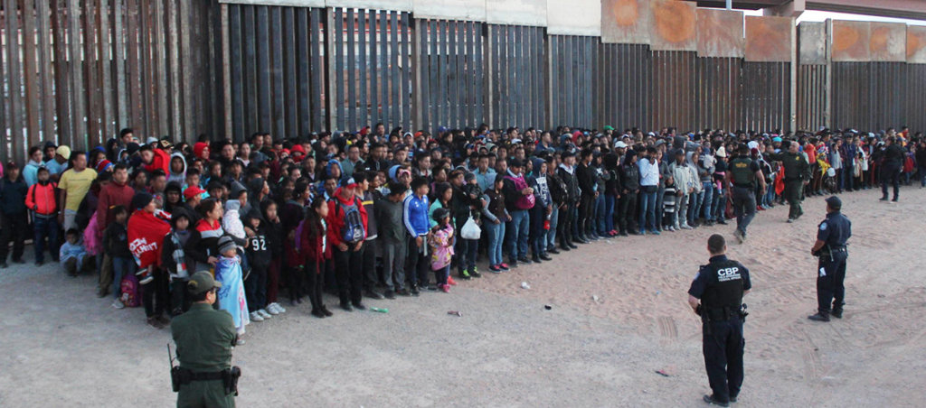 A photo of at least 100 youths lined up against fences with a few armed CBP officers.