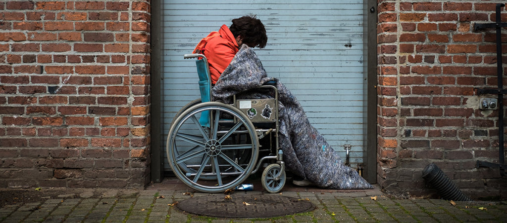 A photo of an unhoused person in a wheelchair and a sleeping bag outside.