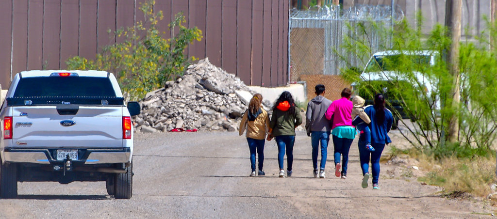 A photo of six people walking to the right and a car to the left.